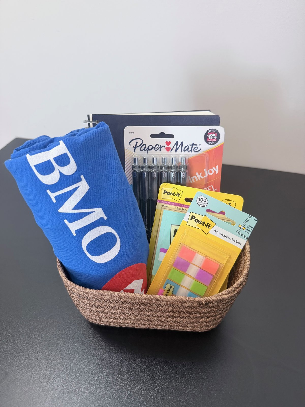 Corporate welcome basket with a company-branded t-shirt, notebook, pens, and sticky notes in a woven basket